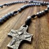 Silberner Rosenkranz liegt auf dunklen Holztisch - das Kreuz ist im Fokus. Auf dem Kreuz sind Brot und Wein zu sehen