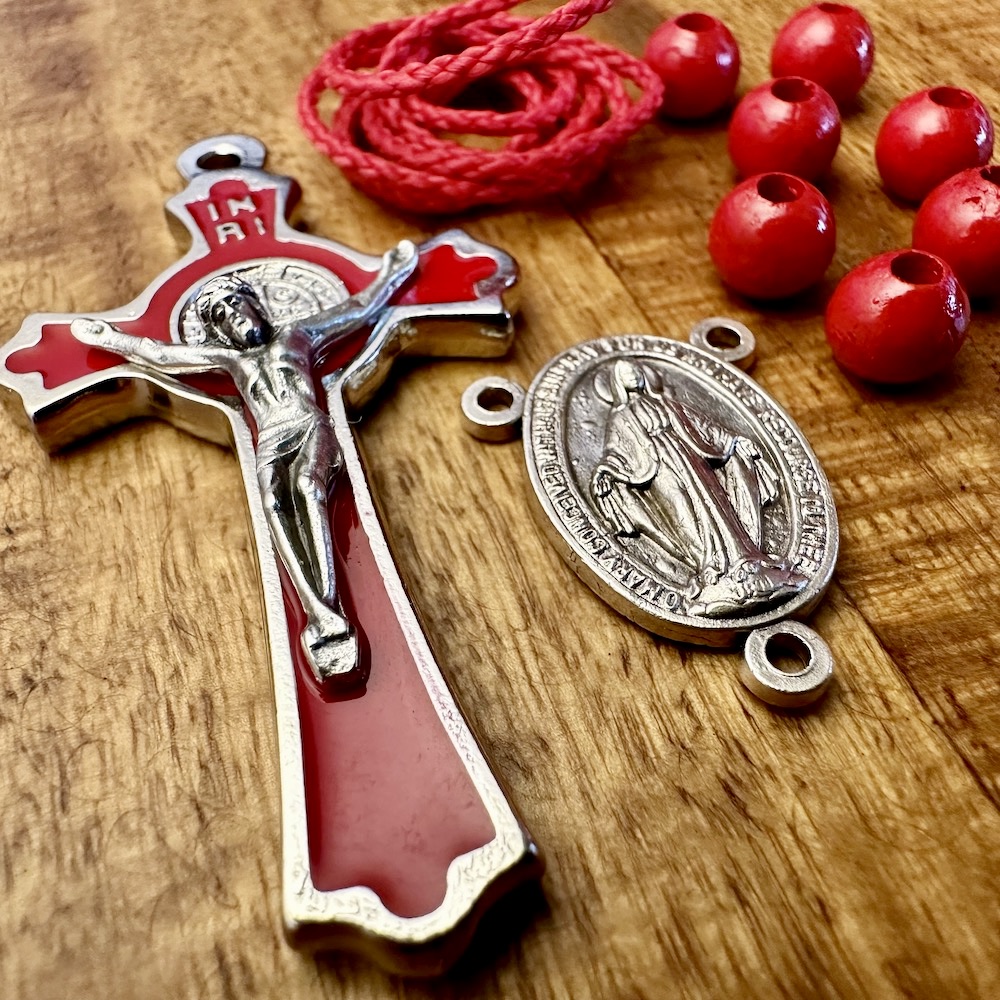 Es liegt ein rotes Kreuz, eine wundertätige Medaille, ein Stück rote Kordel und einige rote Holzperlen auf einem dunklen Holztisch