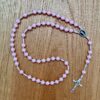 Ein heller rosa Rosenkranz liegt auf einem Holzuntergrund. Als Mittelstück sieht man den barmherzigen Jesus.