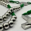 Rosenkranz mit 6mm Metallperlen und grüner Kordel liegt auf weisen Hintergrund - Nahaufnahme einiger Perlen und ein Teil des Kreuzes