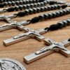 Es liegen offene 10er Rosenkränze auf einem Holzuntergrund. Man sieht einige Benediktuskreuze und schwarze Hämatitperlen