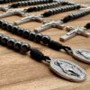 Man sieht eine wundertätige Medaille eines offenen 10er Rosenkranzes mit Hämatit Perlen und schwarzer Kordel - im Hintergrund sind noch weitere 10er mit Benediktuskreuz zu sehen.